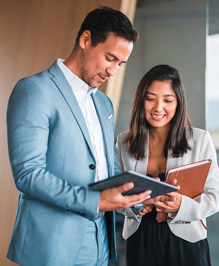 Two business professionals stand together, looking at a tablet. The man wears a light blue suit; the woman holds a notebook and wears a white blazer over a black top.